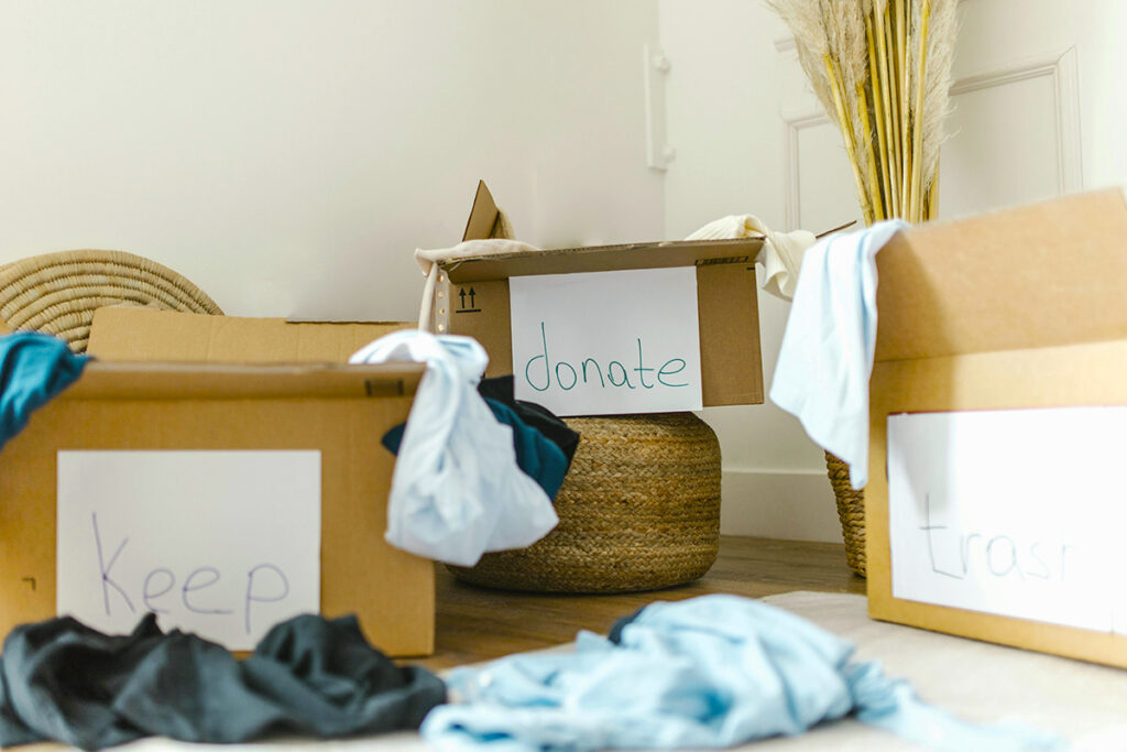 Cardboard boxes labeled 'keep,' 'donate,' and 'trash' on the floor of a room with clothing items scattered about.