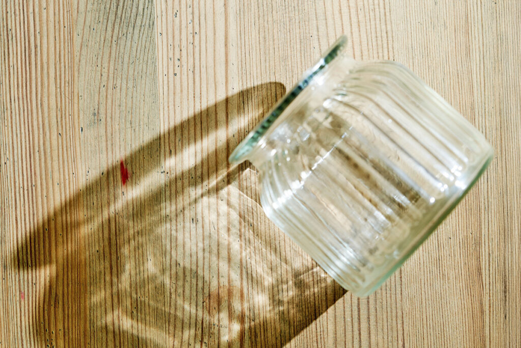 An empty, fluted clear glass candle jar creating a light reflection on a wood surface.