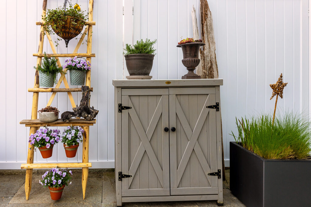 A gray outdoor storage cabinet next to a wooden ladder with hanging pots full of herbs and flowers.