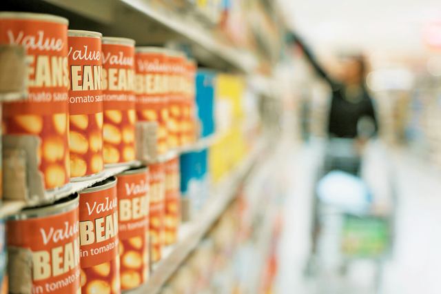 Grocery store aisle showing canned beans in tomato sauce in focus. Down the aisle and out of focus is a woman with a shopping cart.