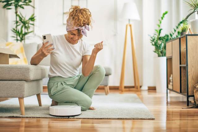 A woman crouched on her living room floor holding a smartphone and smiling at a robot vacuum that is cleaning an area rug.