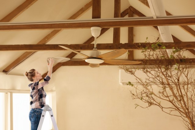 Woman on a ladder using a pillowcase to clean the blades of a ceiling fan. The ceilings are high and vaulted with exposed wood beams.