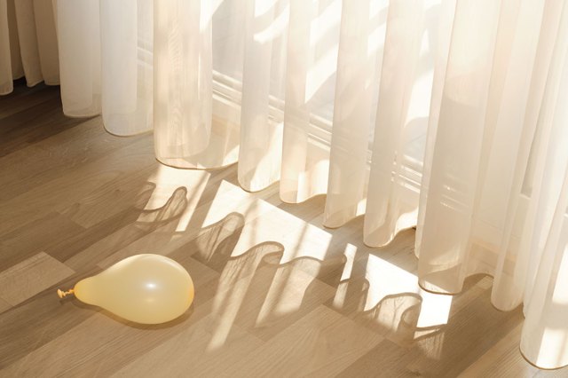 A yellow, partially-inflated balloon on a light wood floor next to a slider door covered by sheer curtains. Sunlight is filtered through the curtains and casting onto the floor near the balloon.