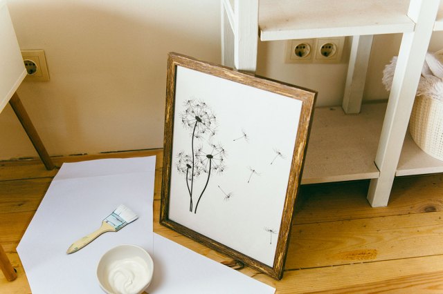 Dried dandelion artwork with a white background in a wood frame. It's perched on craft paper with a paint brush and a bowl of Mod Podge next to it.