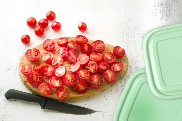 About two-dozen cherry tomatoes sliced in half on a wooden cutting board. Next to them is a small knife and two mint green Tupperware lids.