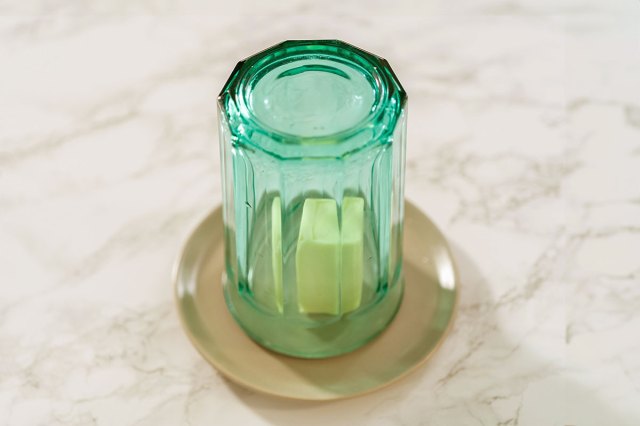 A green drinking glass placed upside down to cover a pat of butter that's sitting on a small, beige plate on a marble countertop.