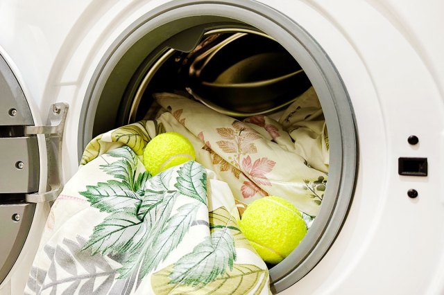 An open front-loading dryer with a patterned comforter spilling out of it. On top of the comforter are two bright yellow tennis balls.