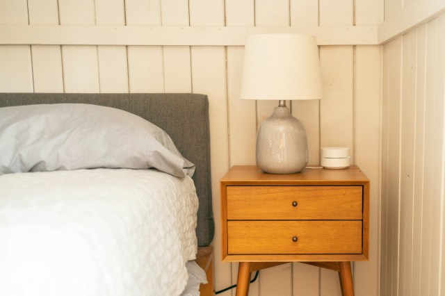 A bed with a gray upholstered headboard next to a wooden mid-century modern nightstand with two drawers. On the countertop is a ceramic table lamp and a smart home device.