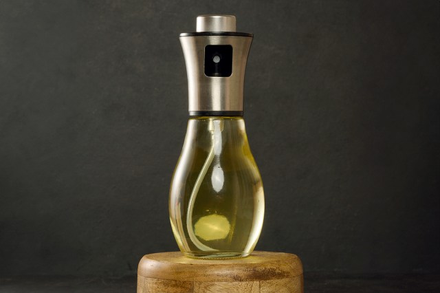 A reusable spray bottle full of cooking spray sitting on a wooden stool against a black background. The spray bottle has a chrome lid and a transparent body.