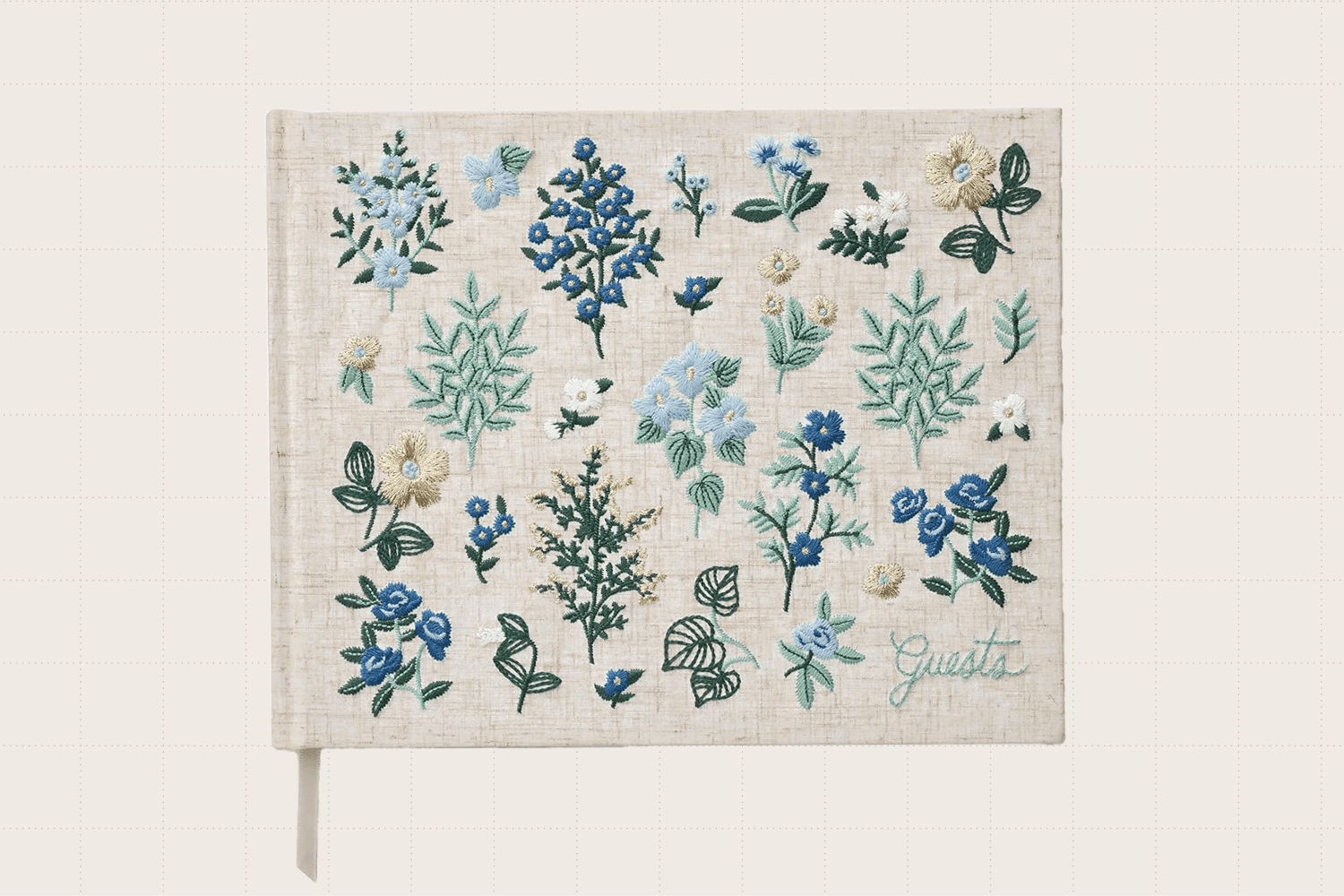 The RIFLE PAPER CO. Wildwood Embroidered Fabric Guest Book. The hard cover is wrapped in burlap-colored cloth with blue, white, and green flowers embroidered on it. In the bottom right corner, it reads 'Guests.' The book has 144 ruled pages.