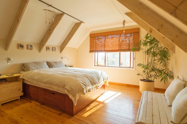 An attic guest bedroom with a pitched ceiling and wooden beams. The room is off-white, with matching white bedding, a large potted plant, and other natural items like wood furniture and a bamboo window shade.