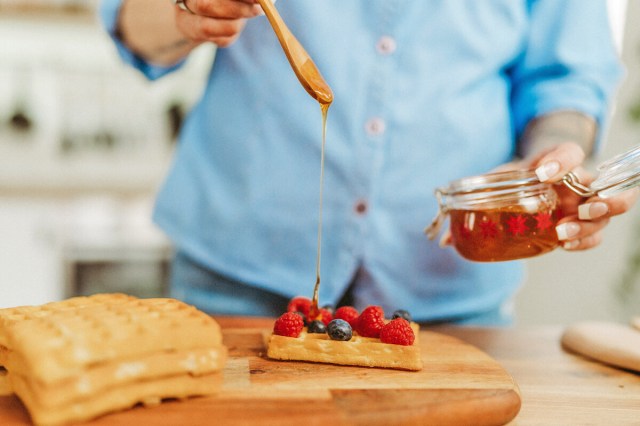 Anonymous person drizzling honey over Belgian waffles topped with blueberries and raspberries. The honey is in an airtight jar and she is using a wooden spoon to add it to the waffles.