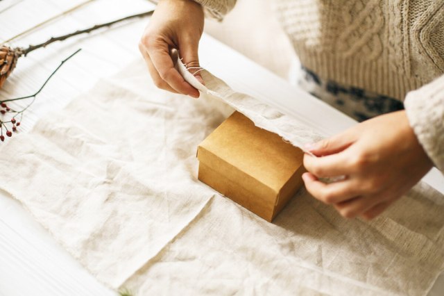 Anonymous person wrapping a small cardboard box in cream-colored cotton fabric. Next to them is a sprig of berries and a pinecone they're presumably going to use for decoration.