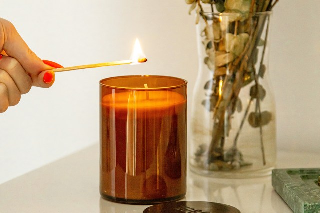 Anonymous person lighting a brand new candle in an amber-colored glass jar using a long, lit match. The candle is on a glass table next to a bouquet of flowers in a clear glass vase.