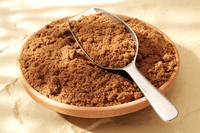 A bowl of brown sugar against a textured surface. In the bowl is a stainless steel scoop that's filled with the ingredient.