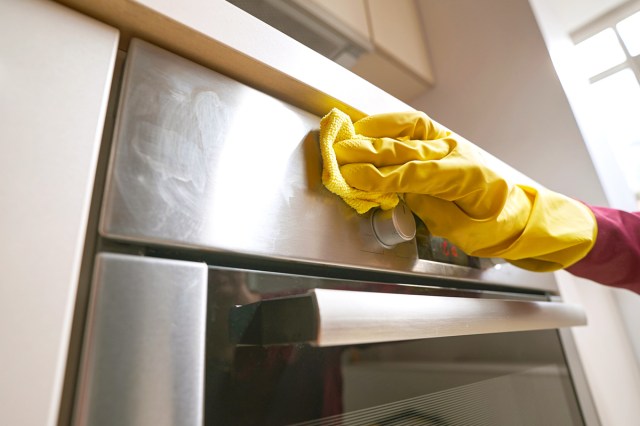 Anonymous person wearing a yellow dishwashing glove cleaning a stainless steel oven with a yellow microfiber cloth.