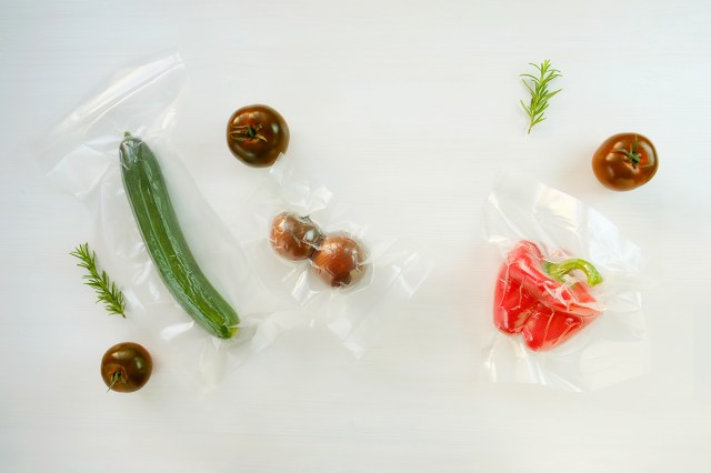 Three types of vacuum-sealed food shot from above, including a cucumber, kumato tomatoes, and a red bell pepper. The vegetables are on a flat, white surface.