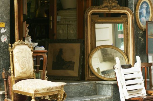 An open doorway of a vintage or thrift store with multiple items positioned near the entrance including two gold framed mirrors, an ornate chair, a painting, and crystal bowls.