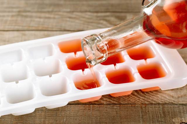 Off-camera person pouring the remains of a red liquid in a clear glass bottle into an ice cube tray that's sitting on a wood countertop.