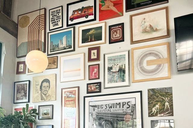 A gallery wall on a large white wall with tall ceilings. The pieces are all framed, different shapes and sizes, and include a mix of photos and artwork.