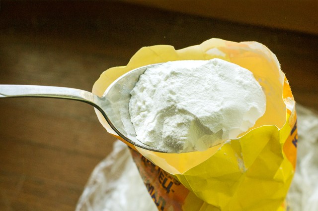 Off-camera person holding a spoonful of cornstarch over a bag of it on a wooden countertop.