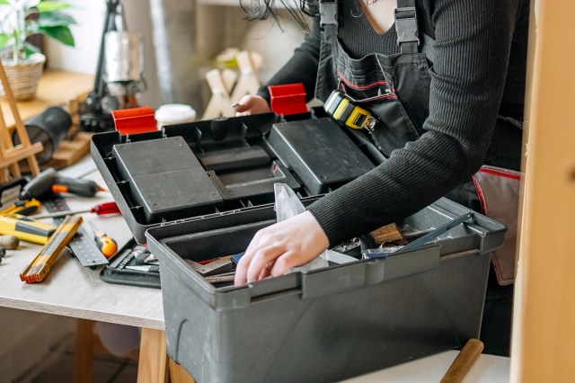 Anonymous person at a work bench with an open toolbox placing something inside of it. She is wearing a work apron and there are tools scattered across the bench she's standing at.