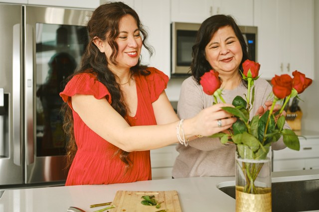 A younger woman wearing red arranging roses in a vase on a kitchen counter. Standing next to her is an older woman who looks to be her mother. She is looking on in approval; both are smiling.