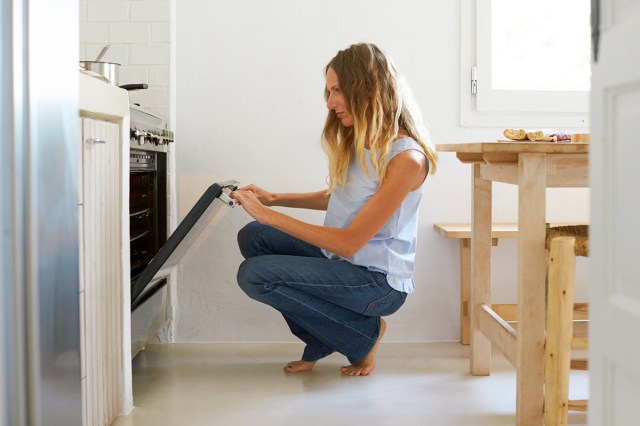 A blonde, middle-aged woman wearing jeans and a sleeveless shirt crouched down in front of an oven, looking at it with the door open.