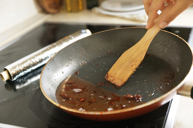 Anonymous person using a wooden spoon to scrape bacon. bits into a pool of grease in a nonstick frying pan on the stovetop. Next to the pan is a roll of aluminum foil.