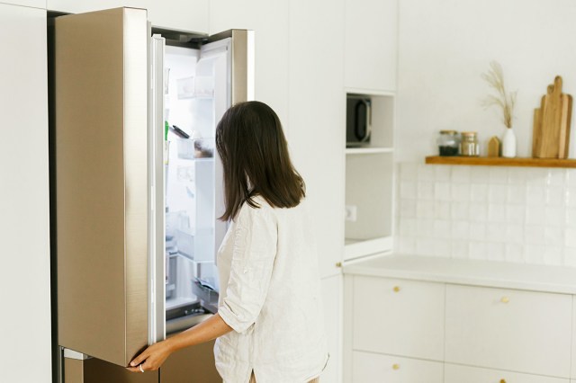 Anonymous woman with dark hair and a white long-sleeve shirt standing at an open refrigerator staring into it. The kitchen is modern-looking and all white.