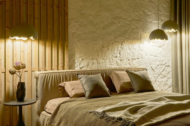 A cozy-looking bedroom featuring a bed with an upholstered headboard, dimly-lit pendant lights, various textured linens on the bed, and a simple vase with dried flowers on the nightstand.