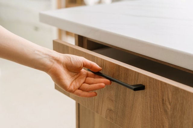 Anonymous person opening a drawer (presumably the junk drawer) in a kitchen. The countertop is white marble, the cabinet is wood, and the hardware is black. The drawer is ajar and the person's hand is on the pull.