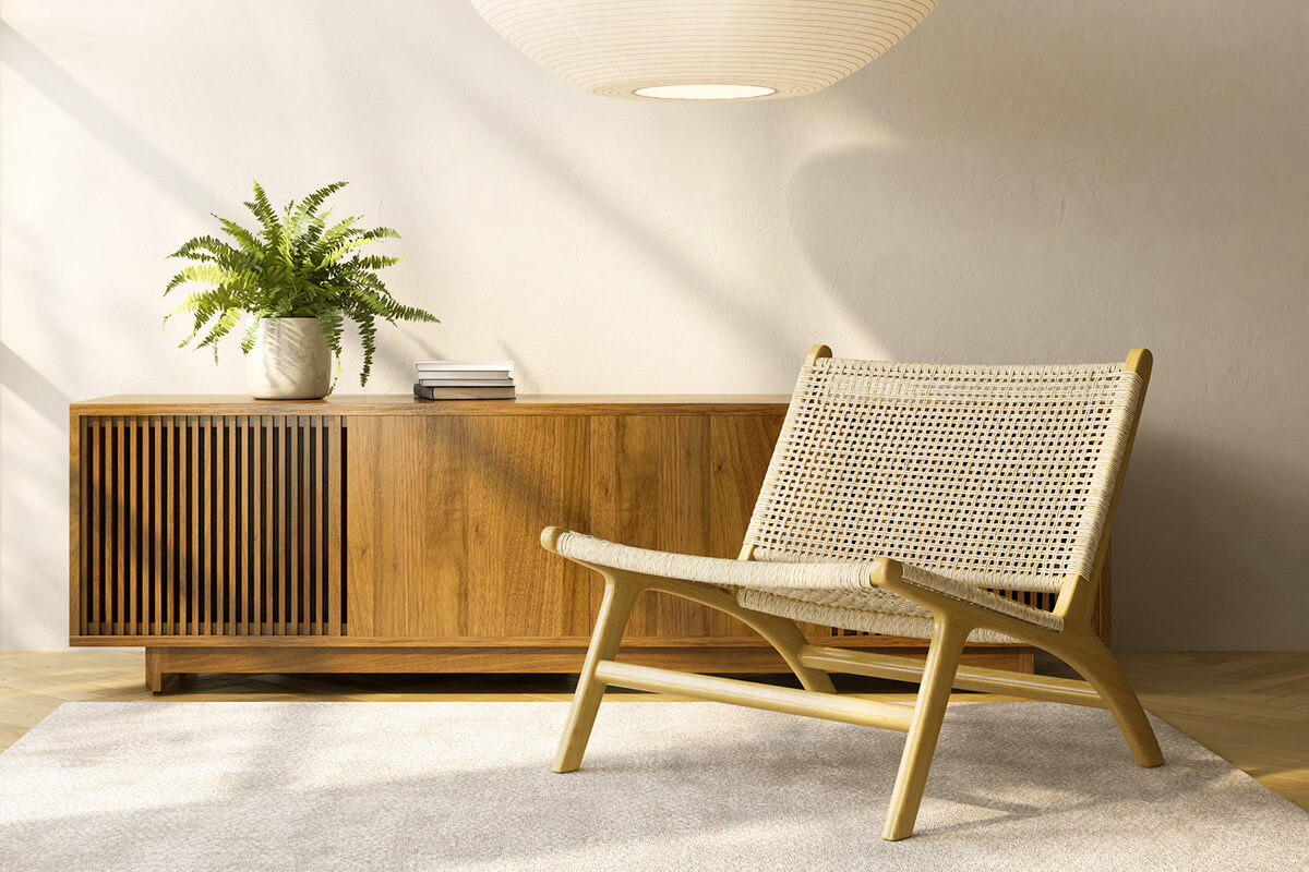 A midcentury modern-style sideboard against a beige wall with a woven, low profile accent chair in the foreground. Underneath the chair is an off-white area rug, and there's a potted fern and three stacked books on the sideboard.