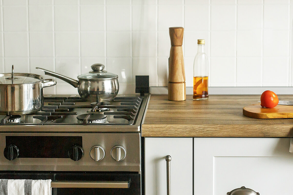 A close-up of a kitchen with a gas stove, wooden countertops, and white cabinetry. On the stove are two stainless steel pots and on the counter is a pepper mill, a bottle of cooking oil, a wooden cutting board, and a tomato.