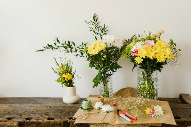 A rustic wooden table with three vases of floral arrangements. In front of the vases is kraft paper, cut blooms, twine, and floral shears.