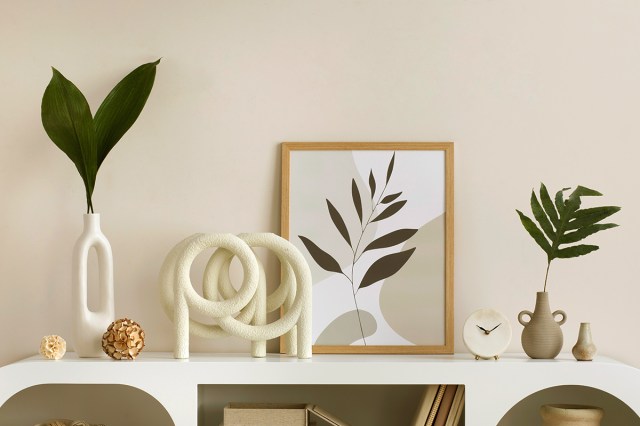 A white, modern, low console table topped with decorative objects including a vase, a sculpture, a framed print, a clock, and two ceramic vases. The console is set against a beige wall.