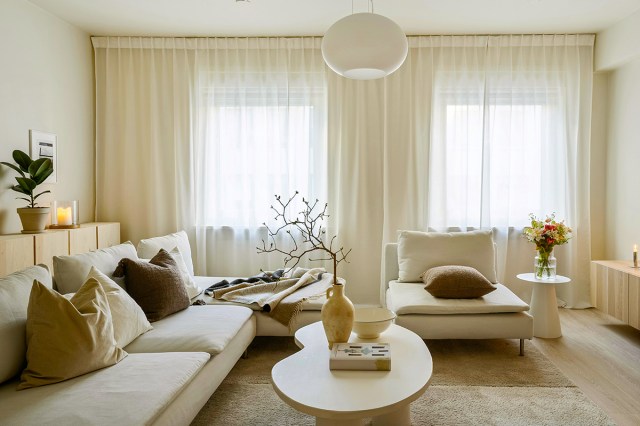 A mostly white and beige living room with a kidney bean shaped coffee table featuring a vignette with a tall vase, ceramic bowl, and a book. There is a large white sectional and consoles on two of the walls.